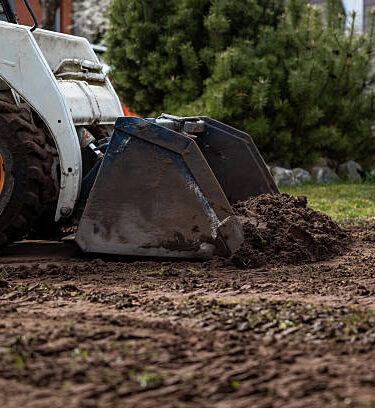 A skid steer loader with orange wheel rims moves soil with its bucket. Nearby are a green bush, a building with horizontal siding, and excavated ground.